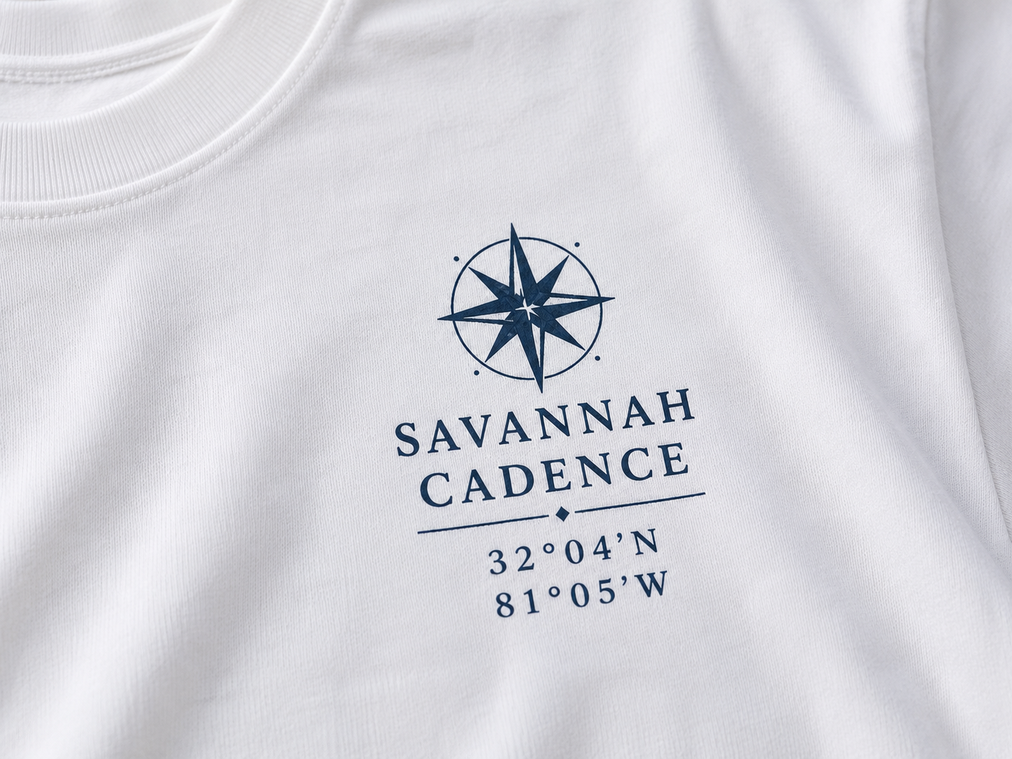Close-up of the Savannah Cadence compass logo on the front left chest of a white Next Level 6210 t-shirt.